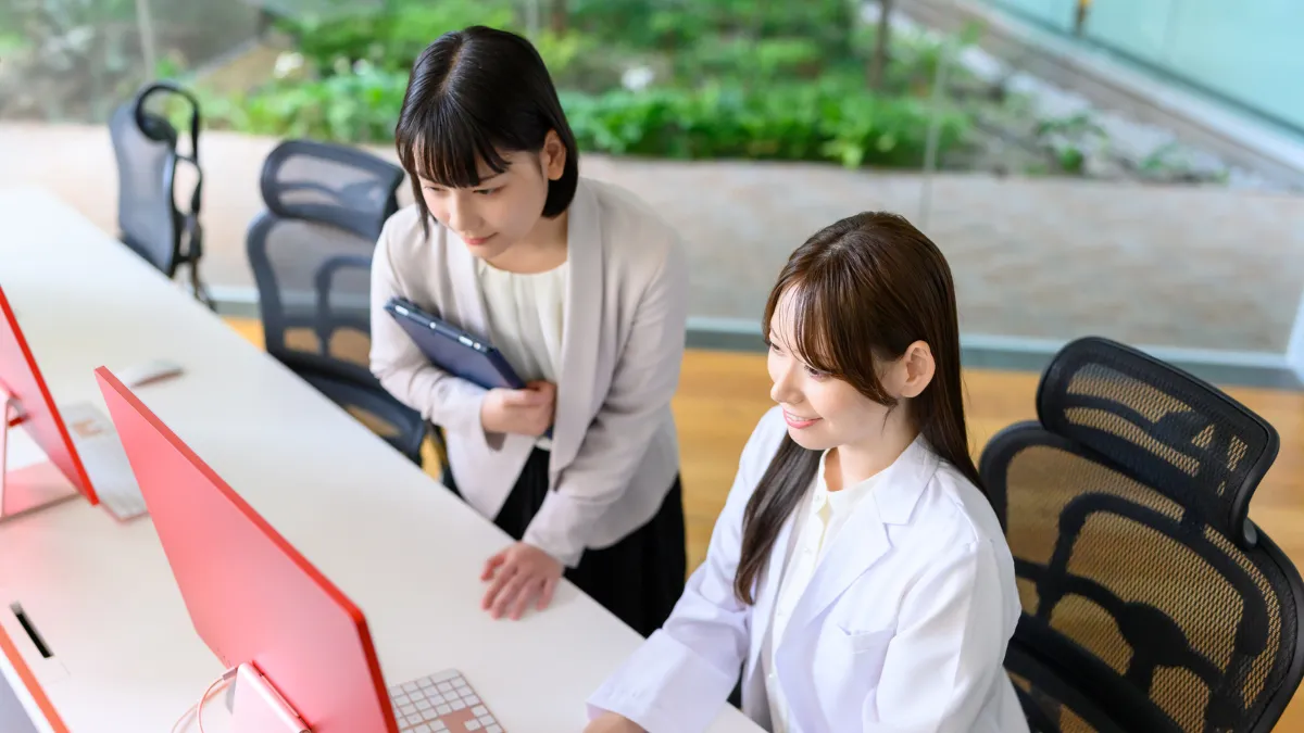 白衣を着た女性とスーツを着た女性がパソコンの前で座って話をしている