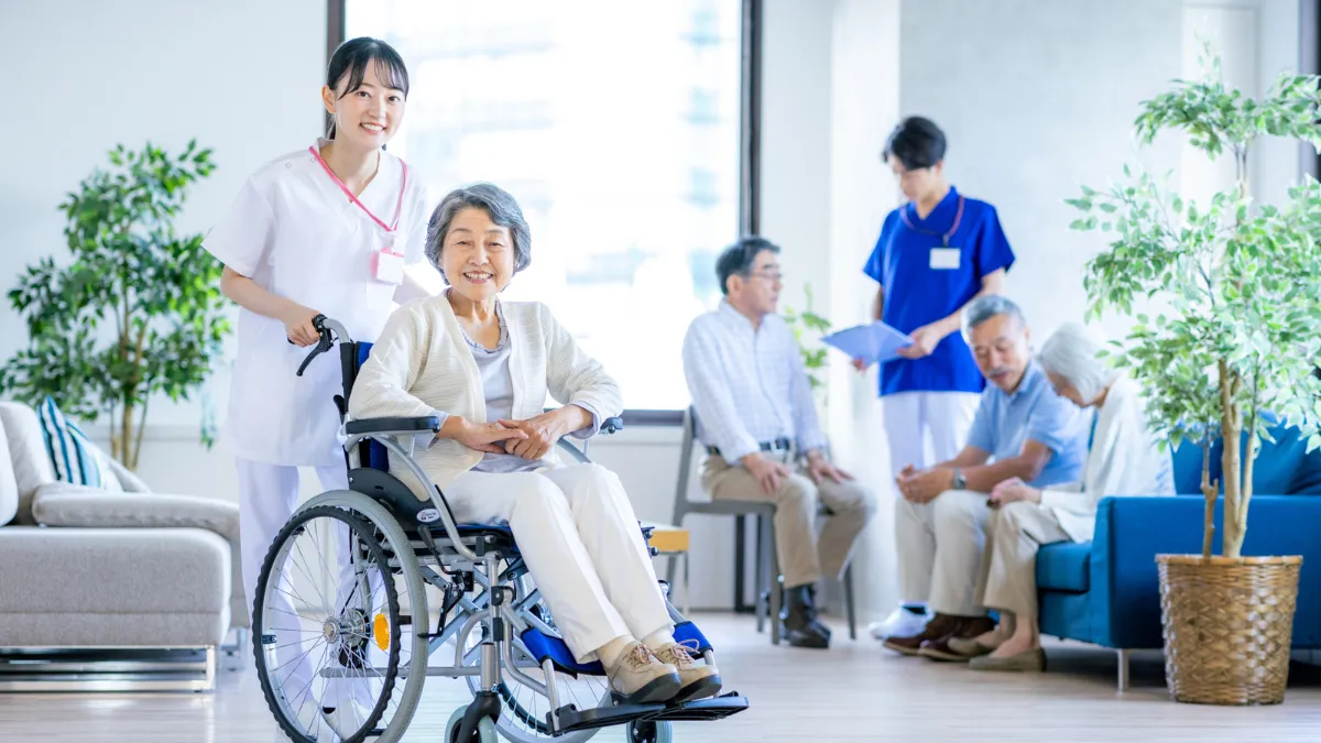 車いすを押してる白衣を着た女性と後ろの方にソファーやいすに座ってる人がいる